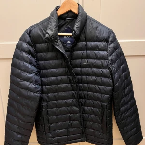 Svart pufferjacka från GANT - Snygg svart pufferjacka från GANT i storlek S. Jackan har en quiltad design med dragkedja framtill och en hög krage för extra värme. 