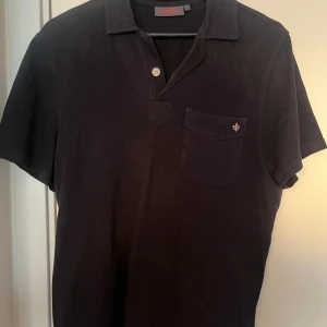 Mörkblå piké från Morris. - Snygg mörkblå piké från Morris med korta ärmar och en stilren bröstficka. Piken har en liten broderad detalj på fickan och en klassisk krage med knappar. Perfekt för en avslappnad stil. Hör av dig vid fråga.