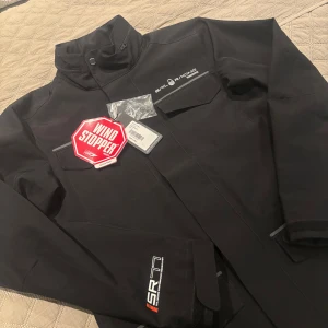 Svart jacka från Sail Racing - Säljer en svart jacka från Sail Racing med Windstopper-teknologi. Jackan har en hög krage och flera fickor med lock. Perfekt för blåsiga dagar. Märket SR-71 syns på ärmen.