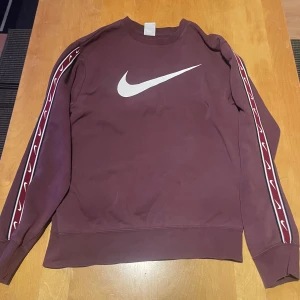 Vinröd sweatshirt från Nike - Säljer en vinröd sweatshirt från Nike med det klassiska vita swoosh-loggan på bröstet. Tröjan har långa ärmar med ett mönster längs ärmarna. Perfekt för en avslappnad stil.
