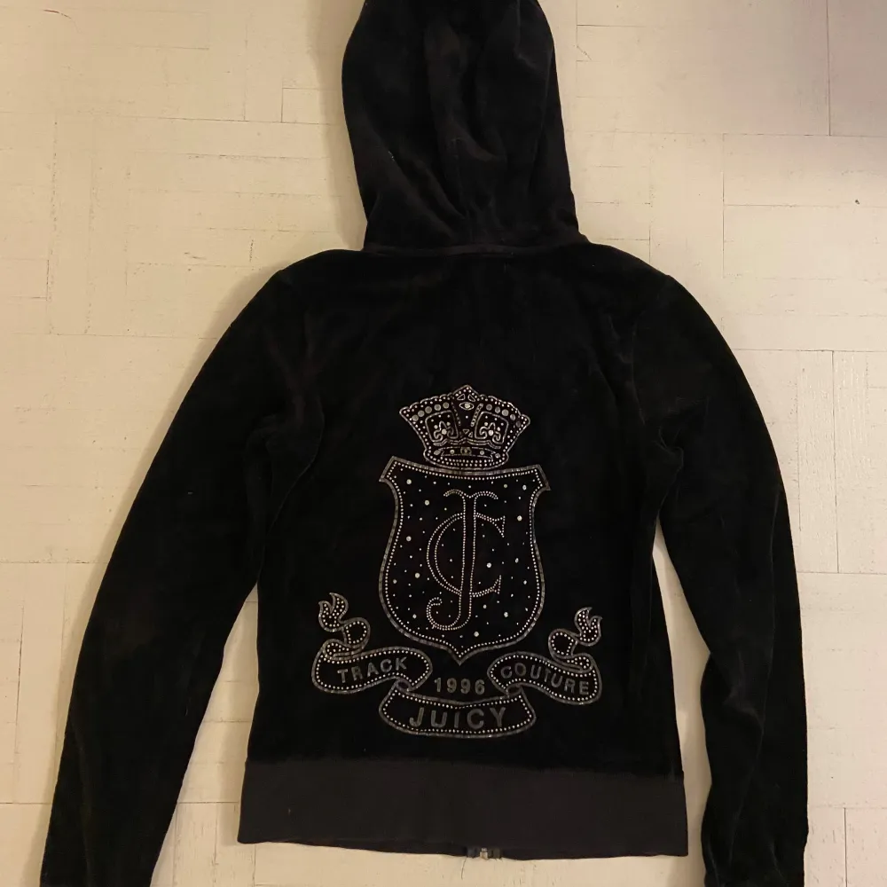 Snygg svart velourjacka från Juicy Couture med huva och dragkedja med ett J på. Jackan har ett glittrigt emblem på ryggen och två fickor fram. Fint skick.. Neuletakit & Villapaidat.