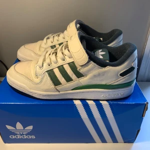 Vita sneakers från Adidas - Snygga vita sneakers från Adidas med gröna detaljer och kardborreband. Skorna har klassiska tre ränder på sidan och en bekväm passform. Perfekta för en sportig look.