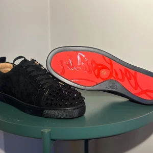 Christian Louboutin - Snygga svarta sneakers i mocka från Christian Louboutin med ikoniska röda sulor. Skorna har svarta nitar på tån och snörning för en edgy look. Perfekta för att ge din outfit en lyxig touch.