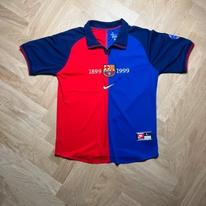 Röd och blå barcleona fotbollströja från Nike - Säljer en klassisk röd och blå fotbollströja från Nike med klubbmärke och årtalen 1899-1999 på framsidan. Tröjan har korta ärmar och en krage med röd och blå rand. Perfekt för samlare eller fans!