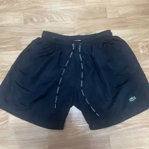 Säljer ett par svarta shorts från Lacoste. De är i nyskick och har en normal passform. Shortsen har en elastisk midja med snörning och är perfekta för sommar och träning. Materialet känns som polyester och de har den klassiska Lacoste-loggan på ena benet.