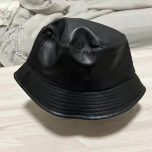 Säljer en snygg och trendig svart bucket hat i skinn. Perfekt för att ge din outfit en cool touch. Hatten har en normal passform och är tillverkad i ett mjukt och bekvämt material. Passar både till vardags och fest!