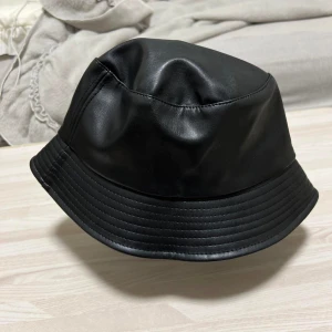 Svart bucket hat i skinn - Säljer en snygg och trendig svart bucket hat i skinn. Perfekt för att ge din outfit en cool touch. Hatten har en normal passform och är tillverkad i ett mjukt och bekvämt material. Passar både till vardags och fest!