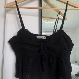 Svart linne från Postie - Säljer ett svart linne i storlek M, men liten i storlek så säger S o har justerbara band så passar de flesta. Det är ärmlöst med smala axelband och har en söt knytning framtill. Materialet är lätt och luftigt, perfekt för sommardagar. Toppen har även fina spetsdetaljer som ger den en extra touch.