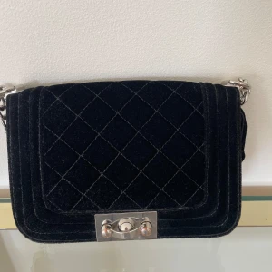 Svart quiltad axelväska med kedja - Säljer en snygg svart axelväska i quiltad design. Väskan är gjord av sammet och har en silverfärgad kedja som axelrem. Den har en stilren och elegant look med en metallspänne framtill. Perfekt för både vardag och fest!