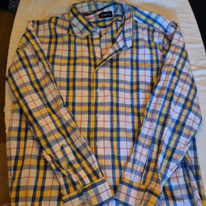 Rutig skjorta från OHMYS - Säljer en snygg rutig skjorta från OHMYS i storlek XL. Skjortan har ett färgglatt mönster i blått, gult och vitt med inslag av rött. Den är långärmad och har knappar längs hela framsidan. Perfekt för både vardag och fest!