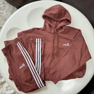 Säljer en snygg och bekväm brun hoodie och matchande byxor från Adidas. Hoodien har en klassisk design med en stor ficka fram och Adidas-loggan på bröstet & ryggen. Byxorna har de ikoniska tre ränderna längs sidorna och en liten Adidas-logga på ena benet. Perfekt set för både träning och vardag! Använt vid enstaka tillfällen, så gott som nytt! 