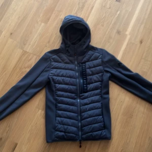 Svart pufferjacka från Zavetti Canada - Säljer en svart pufferjacka från Zavetti Canada. Jackan har en huva och dragkedja framtill. Den är vadderad och perfekt för kyligare väder. Jackan är i storlek S och passar bra för höst och vår.