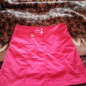Rosa kortkjol med knappar - Säljer en superfin rosa kortkjol shorts som är perfekt för sommaren! Kjolen har två knappar fram och en liten dekorativ knappdetalj. Den är gjord av ett bekvämt material som känns skönt mot huden. Passar perfekt till både vardag och fest. Storlek är inte angiven men den ser ut att vara i en mindre storlek.stl 40