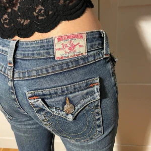 True Religion jeans - Säljer ett par snygga True Religion jeans i storlek S. De är blåa med slitningar på framsidan och har en låg midja. Jeansen är bootcut. Bakfickorna har coola detaljer med knappar och broderi.