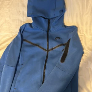Nike Tech - Säljer en snygg blå hoodie från Nike. Den har en dragkedja framtill och en ficka med dragkedja på ärmen. Perfekt för både träning och vardagsbruk. Hoodien är i bra skick och väldigt bekväm att ha på sig. Passar perfekt för höst och vår.