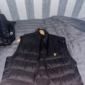 Svart dunväst från Lyle & Scott - Säljer en snygg svart dunväst från Lyle & Scott. Västen är quiltad och har en dragkedja framtill. Den är perfekt för höst och vinter, och håller dig varm och bekväm. Västen är i nyskick och har en klassisk stil som passar till det mesta. Perfekt för lager-på-lager-look eller som ytterplagg under mildare dagar.