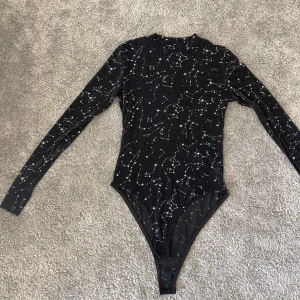 Svart glittrig bodysuit - Säljer en superfin svart bodysuit med glittrigt stjärnmönster. Den är långärmad och gjord i ett stretchigt material av 95% polyester och 5% elastan. Perfekt för festliga tillfällen när du vill glänsa lite extra! ✨