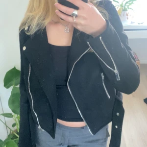 Svart skinnjacka från NLY Trend - Säljer en snygg svart skinnjacka från NLY Trend i storlek 34. Jackan har en cool bikerstil med dragkedjor och knappar i silver. Den är croppad och har ett bälte i midjan för extra stilpoäng. Perfekt för hösten.