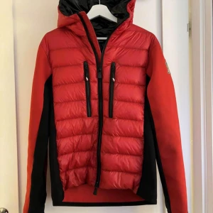 Röd dunjacka från Moncler - Säljer en snygg röd dunjacka från Moncler. Jackan har en cool design med svarta detaljer och en logga på ärmen. Den är perfekt för kalla dagar med sin varma dunfyllning. Jackan har en dragkedja framtill och två praktiska fickor med dragkedjor. Perfekt för vintern! ❄️