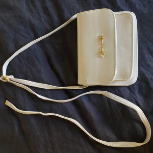 Elegant vit axelväska med gulddetaljer - Säljer en superfin vit axelväska. Den har en stilren design med en smal axelrem och snygga gulddetaljer på framsidan. Väskan har flera fack inuti, perfekt för att organisera dina saker. Den passar perfekt till både vardag och fest🕺