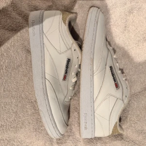 Vita Reebok sneakers med rutigt mönster - Säljer ett par vita Reebok sneakers i klassisk stil. Skorna har ett snyggt rutigt mönster på hälen och insidan, vilket ger dem en unik touch. De är i skinn och har snörning framtill. Perfekta för vardagsbruk och passar till det mesta i garderoben. Skorna är välanvända men fortfarande i bra skick.