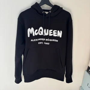 Svart hoodie från Alexander McQueen - Säljer en svart hoodie från Alexander McQueen med stor vit logga på framsidan. Hoodien har en klassisk design med dragsko i huvan och en stor magficka. Perfekt för en stilren och bekväm look. Köpt av leverantör alltså finns inga kvitton eller og.