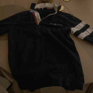 Marinblå pikétröja från Sail Racing - Snygg marinblå pikétröja från Sail Racing med vita och röda detaljer. Tröjan har en broderad logga på bröstet och en stor design på ryggen. Perfekt för en sportig look!