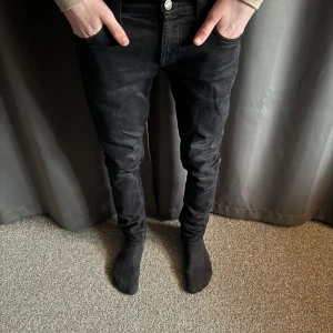 Svarta jeans från Nudie Jeans - Säljer ett par svarta jeans från Nudie Jeans. De har en slim passform och är tillverkade i ett bekvämt denimtyg. Klassisk design med fem fickor och knappgylf. Perfekta för en stilren look. Moddelen är 177cm, bara att skriva vid funderingar