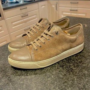 Bruna sneakers från Lanvin - Snygga bruna sneakers från Lanvin i mocka med skinn på tåpartiet. Skorna har en klassisk snörning och en vit sula som ger en stilren look. Perfekta för en avslappnad stil.