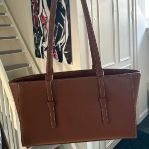 Brun axelväska i skinn - Snygg brun axelväska i skinn med stilrena linjer och vita sömmar. Väskan har långa handtag och en rymlig insida, perfekt för att bära med sig det nödvändigaste. En klassisk design som passar till många tillfällen.