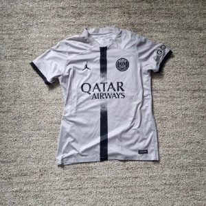 Grå psg tröja Jordan - Säljer en stilren grå fotbollströja från Jordan med svart vertikal rand och PSG-logga på bröstet. Tröjan har korta ärmar med svarta detaljer och Qatar Airways som sponsortryck. Perfekt för fotbollsfans som vill ha något unikt!