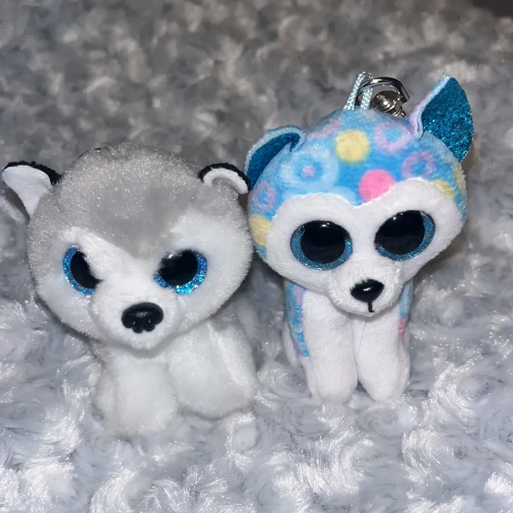 Två små plyschdjur med nyckelringar. Ena är en vit och grå hund med blå glittriga ögon. Den andra är en vit och blå hund med färgglada prickar och blå glittriga ögon.💘Perfekta som accessoarer för väskan eller nycklarna. Nypris 50kr/st💕. Asusteet.