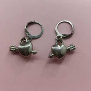 Silverfärgade örhängen med hjärta och pil - Charmiga silverfärgade örhängen med ett hjärta genomborrat av en pil. Perfekta för att ge en romantisk touch till din stil. De har en enkel och säker låsmekanism. Handgjorda.