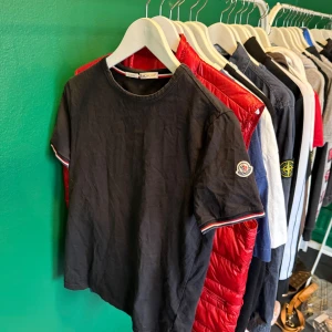 Svart t-shirt från Moncler - Snygg svart t-shirt från Moncler med korta ärmar och broderad logga på ärmen. Den har röd och vit randdetalj vid ärmslut och halsringning. Perfekt för en stilren look.