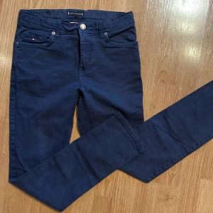 Mörkblå byxor från Tommy Hilfiger - Snygga mörkblå byxor från Tommy Hilfiger med klassisk design. Byxorna har en normal passform och är tillverkade i bomull. De har knappar och dragkedja framtill samt fickor både fram och bak. Storlek 164 Barn