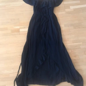 Svart långklänning - Elegant svart långklänning med offshoulder-design och volangdetaljer. Perfekt för en stilfull kväll. Klänningen har en vacker fall och knytband i midjan för en smickrande passform.
