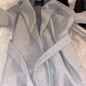 Grå kappa med bälte - Snygg grå kappa med bälte och långa ärmar. Perfekt för kyligare dagar med sin stilrena design och bekväma passform. Ett måste i garderoben för den som älskar klassiska plagg.