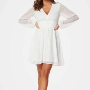 Vit klänning med v-ringning - Super snygg vit klänning med v-ringning och långa, transparenta ärmar. Klänningen har en knytning i midjan och en dragkedjan ryggen. Perfekt inför studenten💗använd 1 gång