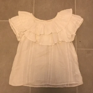 Vit blus från Lindex - Söt vit blus från Lindex med volangdetaljer runt halsen. Blusen har korta ärmar och ett diskret randigt mönster. Perfekt för en somrig look! Inga defekter men har används en del❤️Den har också lite resår i halsen så det går att göra den till off shoulder🌷