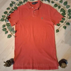 Orange pikétröja från Ralph Lauren - Snygg orange pikétröja från Ralph Lauren med klassisk krage och två knappar. Den har en liten grön logga broderad på bröstet. Perfekt för en stilren och avslappnad look.