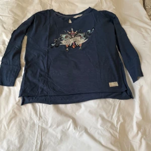 Blå långärmad tröja från Odd Molly - Säljer en blå långärmad tröja från Odd Molly med ett tryck på framsidan. Tröjan är tillverkad i bomull och har en liten rosett på baksidan samt ett litet sprunn i sidorna på tröjan. Tröjan sitter löst och har många fina detaljer.