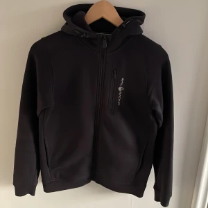 Svart zip från Sail Racing - Säljer en stilren svart hoodie från Peak Performance med dragkedja och huva. Den har långa ärmar och en liten logga på bröstet samt en på huvan. Perfekt för en casual look eller träning.                                                                  Svarar snabbt på frågor!
