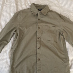 Beige overshirt från H&M - Snygg beige overshirt från H&M med klassisk krage och knappar framtill. Skjortan har en bröstficka och långa ärmar, perfekt för lager på lager-stil. Passar bra till både avslappnade och mer uppklädda tillfällen.