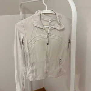 Vit jacka från Lululemon - Säljer en stilren vit jacka från Lululemon med dragkedja framtill och två praktiska fickor med dragkedjor. Jackan har en hög krage och är tillverkad i ett ribbat material som ger en snygg struktur. Perfekt för en aktiv livsstil eller som ett lätt lager under kyligare dagar.