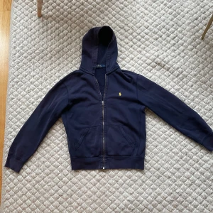 Mörkblå hoodie från Ralph Lauren - Säljer en snygg mörkblå hoodie från Ralph Lauren med dragkedja och det klassiska logotypbroderiet på bröstet. Perfekt för en avslappnad stil. Den har långa ärmar och en bekväm huva.