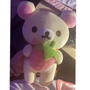Rilakkumma björn med jordgubbe, Gosedjur, plush, ny - Säljer ett gulligt gosedjur i form av en björn med rosa öron och en stor jordgubbe i famnen. Björnen har en prickig rosett runt halsen och är gjord av mjukt material. Perfekt som present eller dekoration!