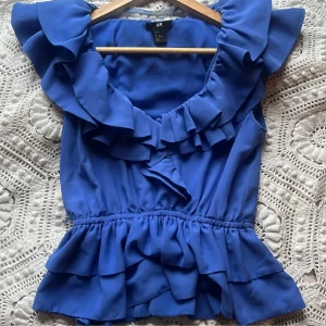 Blå blus från H&M - Säljer en blå blus från H&M med volangdetaljer och v-ringning. Blusen har en elastisk midja och är ärmlös, vilket ger en luftig och bekväm passform. Perfekt för en stilren look.