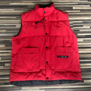 Röd dunväst från Canada Goose - Snygg röd dunväst från Canada Goose med knappar och två stora fickor framtill. Perfekt för kyliga dagar och ger en stilren look. Märkeslogga broderad på bröstet.