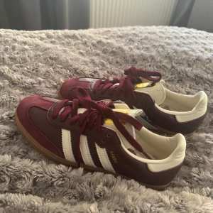 Adidas Samba i vinrött och vitt - Snygga Adidas Samba sneakers i vinrött med vita detaljer och klassiska tre ränder. Skorna har en gummisula och snörning. Helt nya med prislappen kvar.
