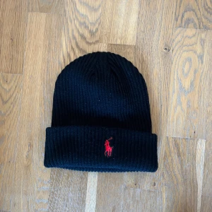 Svart ribbad mössa från Polo - Snygg svart ribbad mössa från Polo med ett rött broderat emblem framtill. Perfekt för att hålla värmen under kyliga dagar. Tillverkad i mjukt material för en bekväm passform.